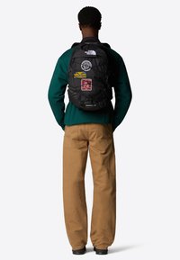 Black backpack with multiple patches on the rear, featuring a quilted design and adjustable straps. Worn over a green sweatshirt and tan pants.