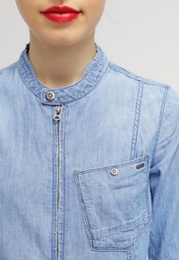 Ljusblå denimjacka med ståkrage, dragkedja fram, bröstficka med tryckknapp och ton-i-ton sömnadsdetaljer.