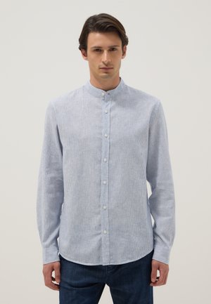 Giovane uomo che indossa una camicia azzurro chiaro a maniche lunghe con collo alla coreana e jeans blu scuro, in piedi davanti a uno sfondo semplice.