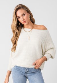 Suéter crema de punto con diseño de acanalado texturizado, que se lleva sin hombros, combinado con jeans de un azul claro y collares dorados superpuestos.