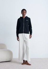Vrouw in zwarte zip-up hoodie en losse witte broek, die blootsvoets op een tapijt staat naast een gebogen, lichtbeige fauteuil.