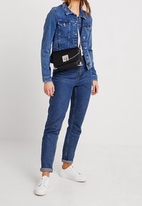 Denimjakke, blå med lommer, kombineret med højtaljede jeans og hvide sneakers. En sort crossbody-taske har en sølvkæde som detalje.