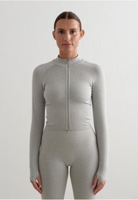 Camiseta ajustada de manga larga gris con cuello alto y cremallera frontal. Textura suave y puños con agujero para el pulgar. Combinada con mallas ajustadas a juego.