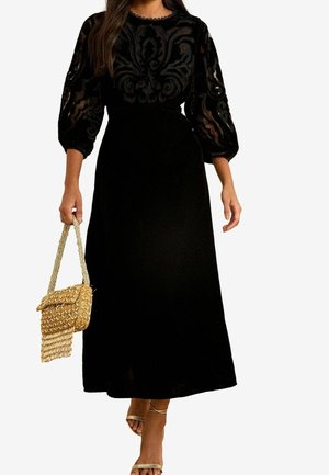Femme portant une robe midi noire avec des manches bouffantes transparentes et à motifs, tenant un sac à main beige tissé et portant des sandales dorées à brides.