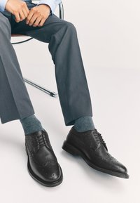 Chaussures brogues en cuir noir à finition texturée, avec des perforations décoratives ; portées avec un pantalon de costume gris et des chaussettes bleues.