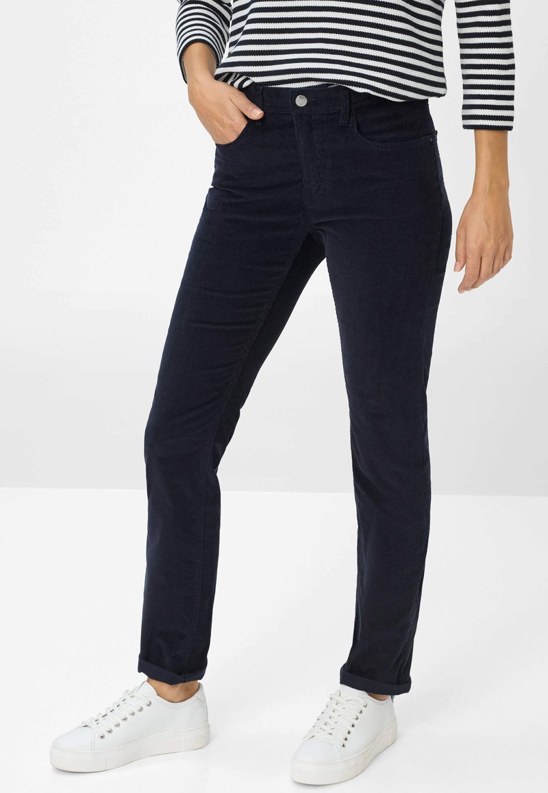 Persoon in slank gesneden donkerblauwe corduroy broek, witte sneakers en een zwart-wit gestreept longsleeve shirt, staat met één hand in de zak.