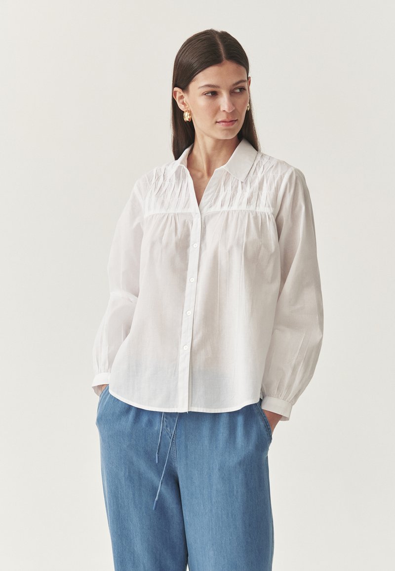 Camicia bianca con colletto, dettagli arricciati sul corpino e maniche lunghe. Abbinata a pantaloni azzurri con cordoncino.