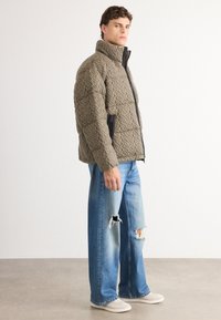 Quilted olivengrøn pufferjakke med høj krave, sort lynlås og tekstureret mønster; parret med slidte, revnede blå jeans og let fodtøj.