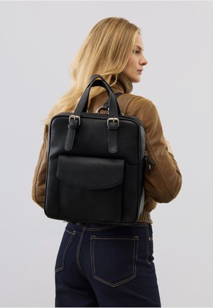 Blonde woman in brown jacket and dark jeans wearing a black leather backpack with front pocket and buckle straps.