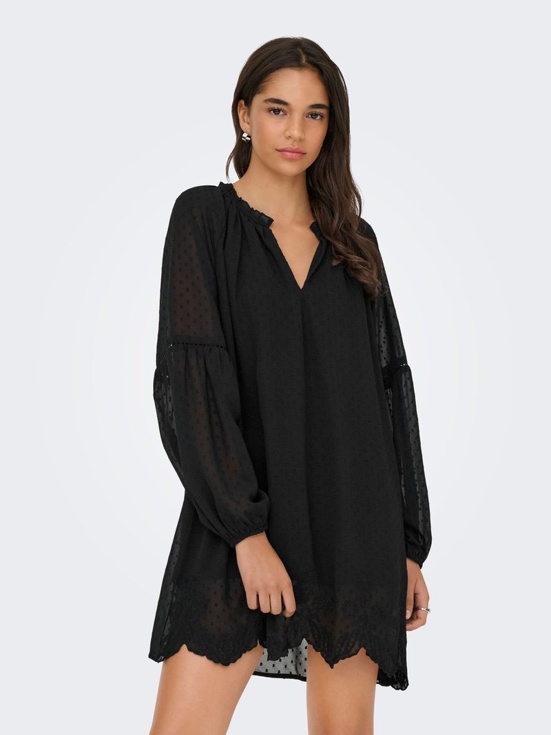 Jeune femme aux longs cheveux foncés portant une robe noire ample avec des manches longues transparentes et un ourlet festonné, debout devant un fond uni.