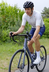 Cycliste sur un vélo de course gris clair, portant un maillot blanc et un short bleu, un casque noir et des chaussures blanches, tenant des poignées de guidon noires.