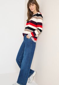 Gestreifter Pullover in Creme, Rot, Schwarz und Braun; kombiniert mit weiten blauen Jeans; akzentuiert durch silberne Sneakers. Strukturierter Strickstoff.