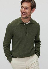 Olivgrön stickad tröja med krage, tre knappar framtill och långa ärmar, tillsammans med ljusbeiga byxor.