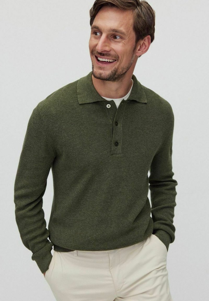 Olivgrön stickad tröja med krage, tre knappar framtill och långa ärmar, tillsammans med ljusbeiga byxor.