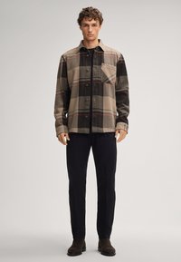 Koszula flanelowa w kratę w odcieniach beżu, czerni i czerwieni; posiada przód na guziki, kieszeń oraz luźny krój, zestawiona z ciemnymi spodniami i brązowymi butami.