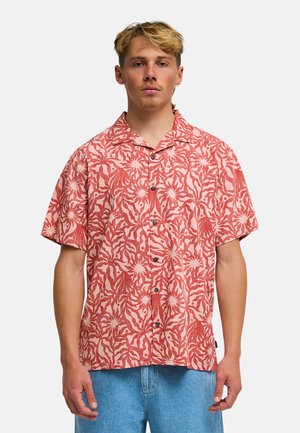 Giovane uomo che indossa una camicia con maniche corte, rossa e bianca con fantasia floreale e jeans azzurri chiari, in piedi davanti a uno sfondo semplice.