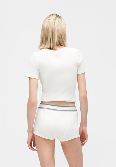 Personne blonde portant un t-shirt blanc court et un short blanc avec une ceinture grise, debout, dos à un fond uni.