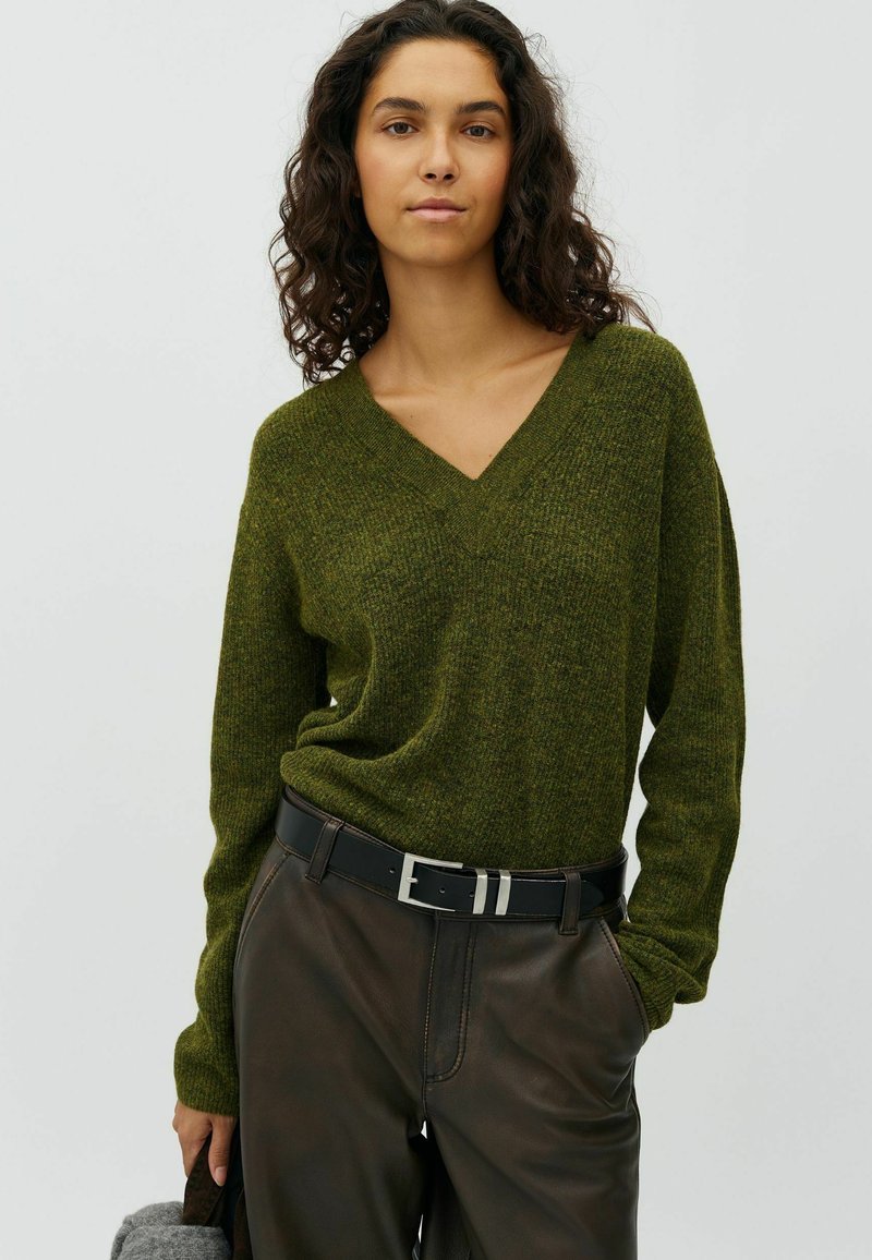 Femme aux cheveux bouclés foncés porte un pull vert à col en V et un pantalon foncé avec une ceinture noire, debout avec une main dans la poche sur fond blanc.