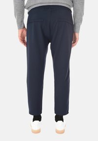 Persona con pantaloni blu navy, maglione grigio, calzini neri e scarpe da ginnastica bianche, in piedi con le mani rilassate ai lati, di spalle rispetto allo spettatore.