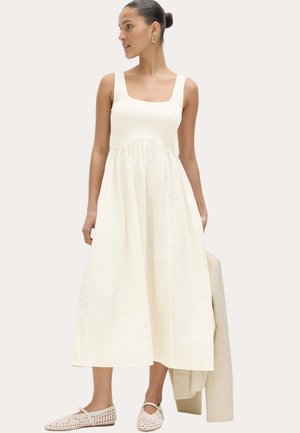 Frau in einem ärmellosen cremefarbenen Midikleid und gewebten cremefarbenen Ballerinas, die eine passende cremefarbene Jacke hält und seitlich vor einem neutralen Hintergrund blickt.