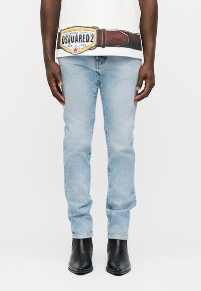 Homme portant un jean slim bleu clair et des bottes noires à bout pointu, debout les mains détendues le long du corps contre un fond blanc.