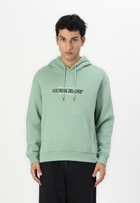 Šviesiai žalia pūkinė striukė su priekinėmis kišenėmis, su raišteliu reguliuojama kapuce ir juodu "Guess Jeans" logotipu ant krūtinės. Pagaminta iš minkštos, tekstūruotos medžiagos.