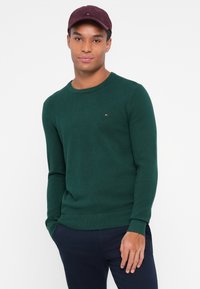 Grüner Strickpullover mit Rundhalsausschnitt, der ein strukturiertes Muster aufweist. Kombiniert mit einer burgunderfarbenen Mütze, vor einem weißen Hintergrund.