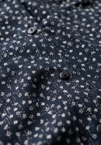 Niebieska koszula z materiału we wzór kwiatowy, z małymi białymi kwiatami, gładką fakturą i czarnymi guzikami.