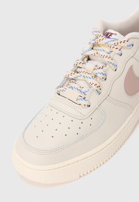 Pogled od blizu na bež Nike tenisku obuću s perforiranim prstom, dušicama u više boja i ružičastim swoosh logotipom sa strane.
