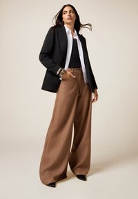Donna che indossa un blazer nero, una camicia bianca, un top nero, pantaloni larghi marroni e scarpe nere con punta, e braccialetti, si trova di fronte a uno sfondo beige.