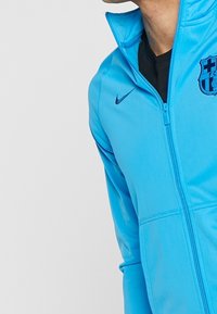 Helleblaue Nike Zip-Jacke mit Barcelona-Logo, glatter Textur, hohem Kragen und kontrastierenden dunklen Details auf der Brust und am Reißverschluss.