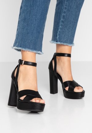 Talons compensés noirs avec des lanières croisées et une bride à la cheville avec une boucle, portés avec un pantalon en jean court à bas effilochés. Texture en cuir lisse.