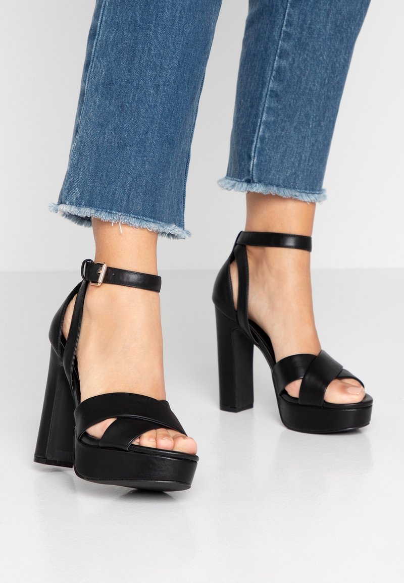 Talons compensés noirs avec des lanières croisées et une bride à la cheville avec une boucle, portés avec un pantalon en jean court à bas effilochés. Texture en cuir lisse.