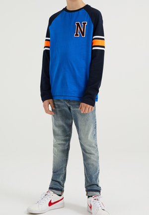 Chemise bleue à manches longues avec manches bleu marine, rayures orange et blanches, et un grand logo "N" sur la poitrine, associée à un jean en denim clair et des baskets blanches.