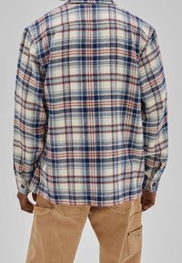 Uomo che indossa una camicia a maniche lunghe a quadri nei toni crema, blu e ruggine, con pantaloni cargo beige, visto da dietro su uno sfondo grigio.