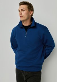 Sudadera azul con cremallera y cuello alto, hecha de una tela suave. Presenta una cremallera de metal y dobladillos y puños acanalados. El modelo lleva pantalones vaqueros negros.