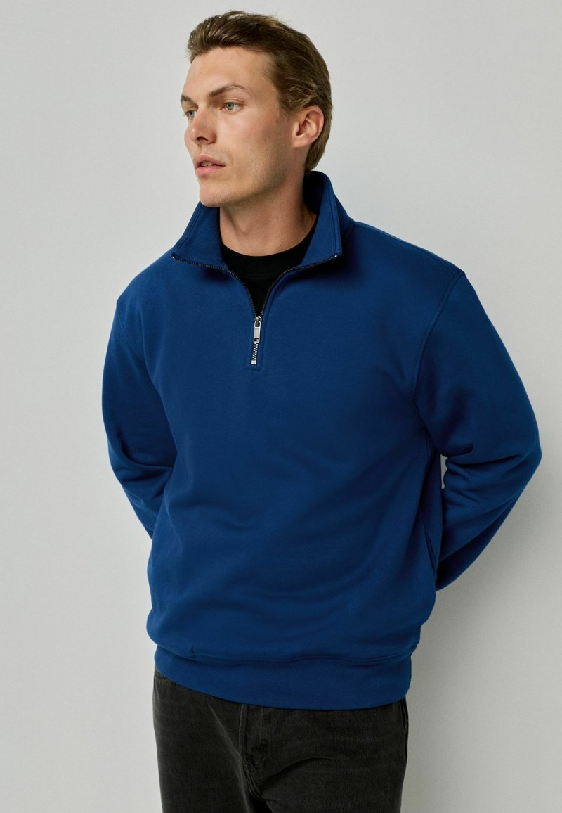 Sudadera azul con cremallera y cuello alto, hecha de una tela suave. Presenta una cremallera de metal y dobladillos y puños acanalados. El modelo lleva pantalones vaqueros negros.