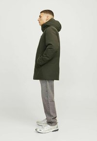 Mørkegrøn parka med hætte, ensartet front og sidelommer. Grå jeans og sølvfarvede sportsko fuldender looket.