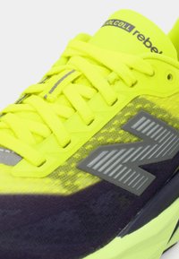 Φωτεινό κίτρινο αθλητικό παπούτσι με επάνω μέρος από δίχτυ, γκρι λεπτομέρειες, υφασμένα κορδόνια και εμφανές γκρι λογότυπο "N" στο πλάι.