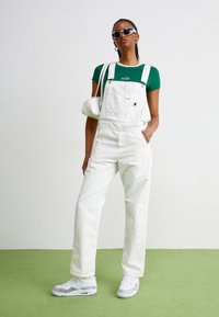 Hvite denimsaloppette med justerbare stropper, sammen med en grønn T-skjorte med hvit tekst. Aksessert med hvit håndveske og sneakers.