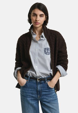 Junge Frau mit kurzem dunklen Haar, die ein gestreiftes Hemd mit Knöpfen, einen dunkelbraunen, grobstrickigen Zip-Pullover, blaue Jeans und einen schwarzen Gürtel trägt, mit den Händen in den Taschen.