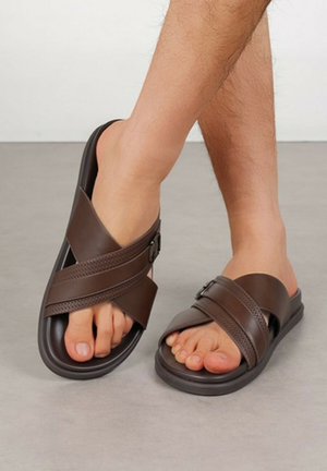 Personne portant des sandales marron foncé sans fermeture avec deux sangles croisées et des boucles, debout sur un sol gris.