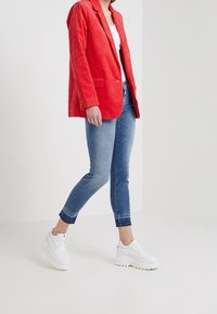 Červená kordurová sako s klopami, spárované se světle modrými skinny džíny a bílými chunky teniskami, zobrazené ve vzpřímené póze.