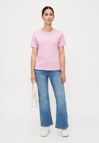 Lys rosa t-skjorte med korte ermer og en dekorativ perlebesetning rundt halsen, kombinert med lyseblå flagrende jeans og hvite sneakers.