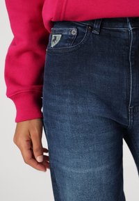 Osoba oblečená v tmavě modrých džínách s malou nášivkou na kapse a v jasně růžovém trikotu s dlouhým rukávem, s rukou uvolněně položenou podél těla.