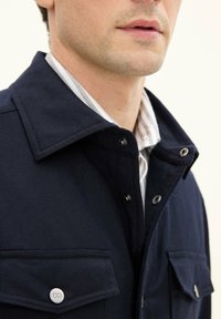 Giacca blu navy con colletto, bottoni a pressione e due tasche frontali. Presenta una consistenza liscia ed è indossata sopra una camicia a righe.