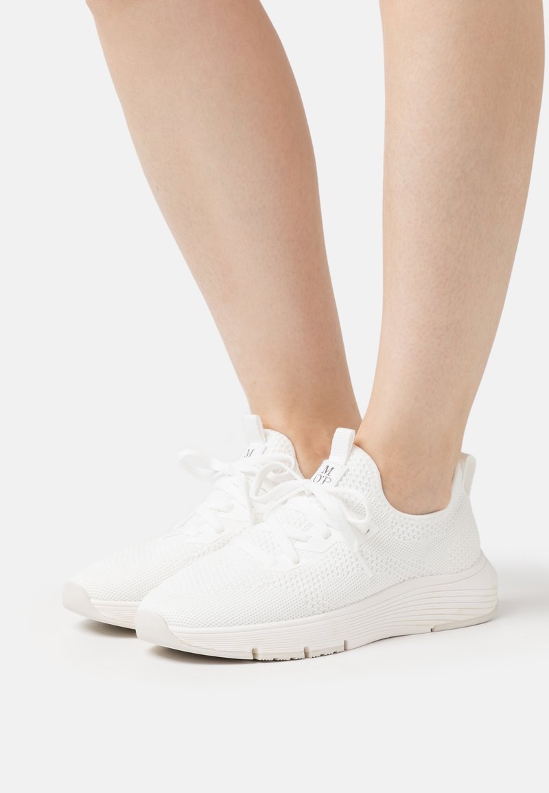 Paire de baskets blanches tricotées portées par une personne avec les jambes et chevilles visibles sur un fond uni.