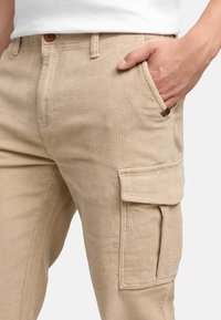 Beige kordfløyelsbukser med ribbet tekstur, som har sidelommer och en knapp i midjan. Enkel design med rak passform.