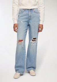 Jasnoniebieskie spodnie dżinsowe z wysokim stanem, o luźnym kroju, z distressed detalami, dużymi przetarciami na kolanach oraz postrzępionymi krawędziami. W zestawie z beżowymi sneakersami.