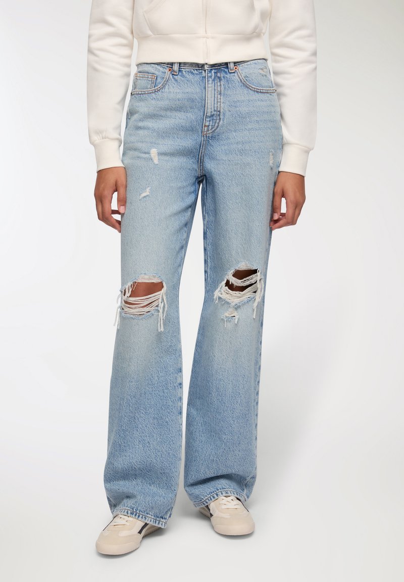 Jasnoniebieskie spodnie dżinsowe z wysokim stanem, o luźnym kroju, z distressed detalami, dużymi przetarciami na kolanach oraz postrzępionymi krawędziami. W zestawie z beżowymi sneakersami.
